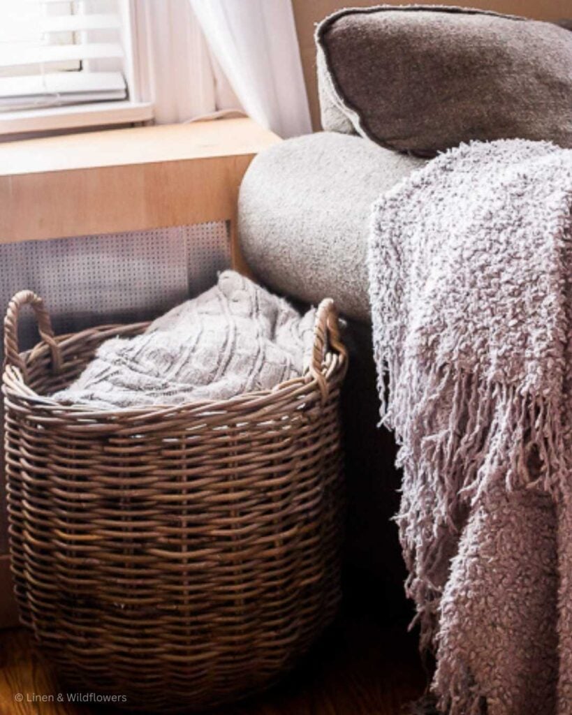 A large wicker basket filled with cozy throw blankets next to a bulky chair with a grey cozy blanket draped over the arm of the chair.