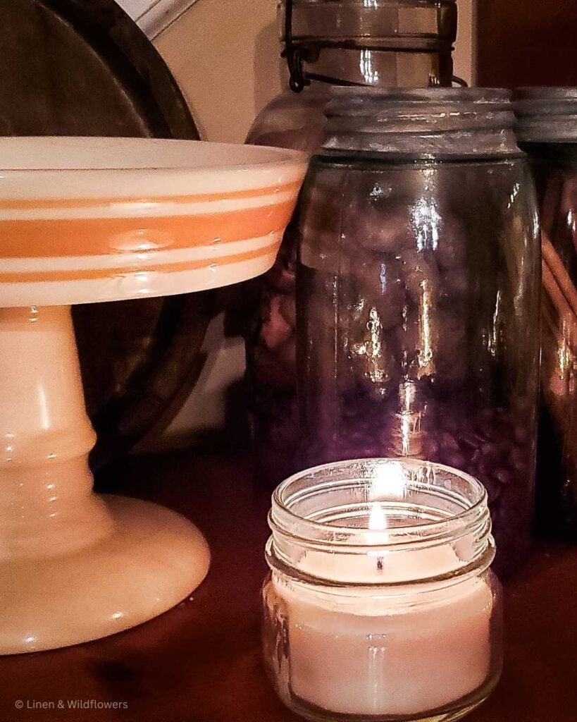 A pedestal dish next to a lit candle and vintage blue mason jars filled with dry goods that set the mood for a cozy atmosphere.