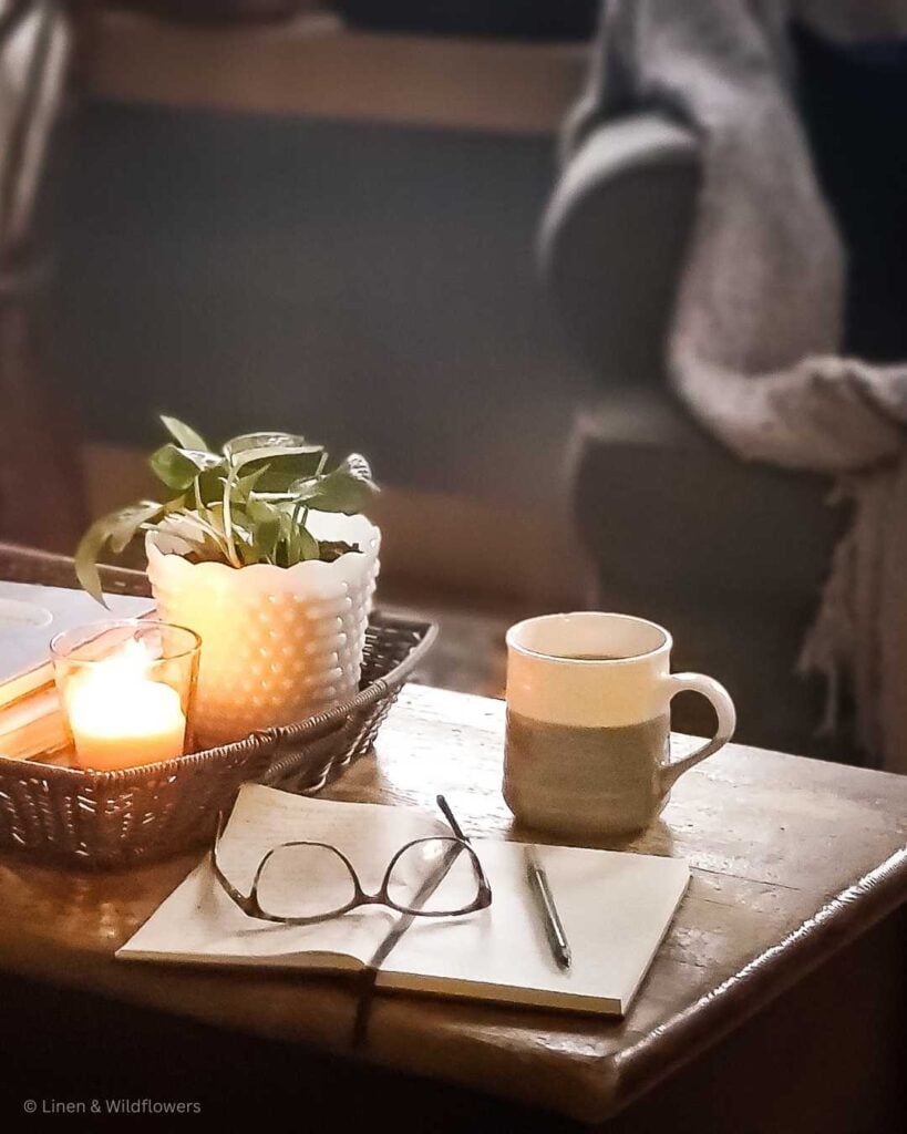 On the blanket chest is a potho plant, hot cup of coffee, a journal, pen and with eye glasses to snuggle up on the couch to write your thoughts on a gloomy day with a lit candle near by.
