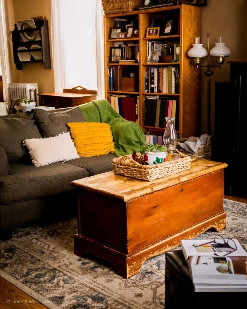 A livingroom that is warm & inviting and has a cozy atmosphere. On the couch is a comfy green throw blanket & throw pillows. On the blanket chest that is used a coffee table has a basket ray witha oil lamp for that ambiance feel, a plant & home decor books for light reading.