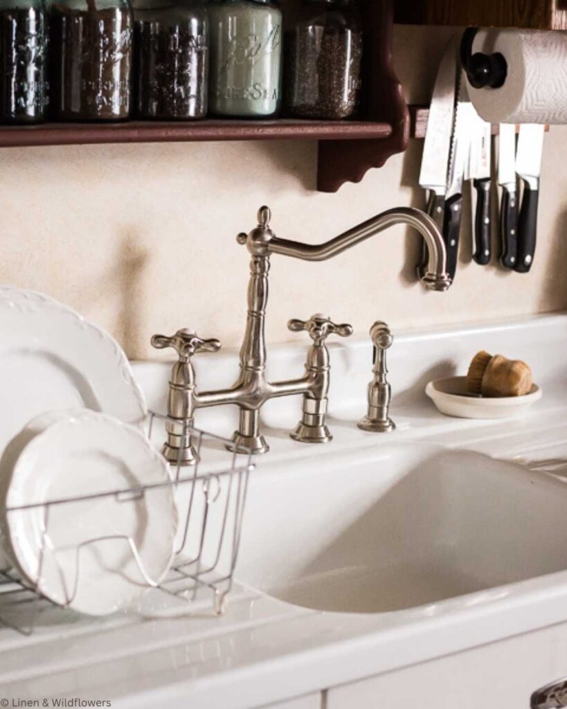 A kitchen counter with a vintage-style silver faucet, dish rack with white plates, mason jars on a shelf, knives in a holder, and a paper towel roll.