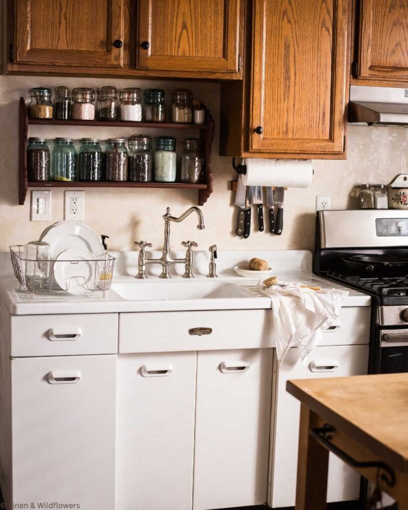 Cozy kitchen with wooden cabinets, a white vintage farmhouse sink with an elegant faucet, and mason jars on a shelf. A dish towel drapes over the counter, evoking a warm, rustic feel.