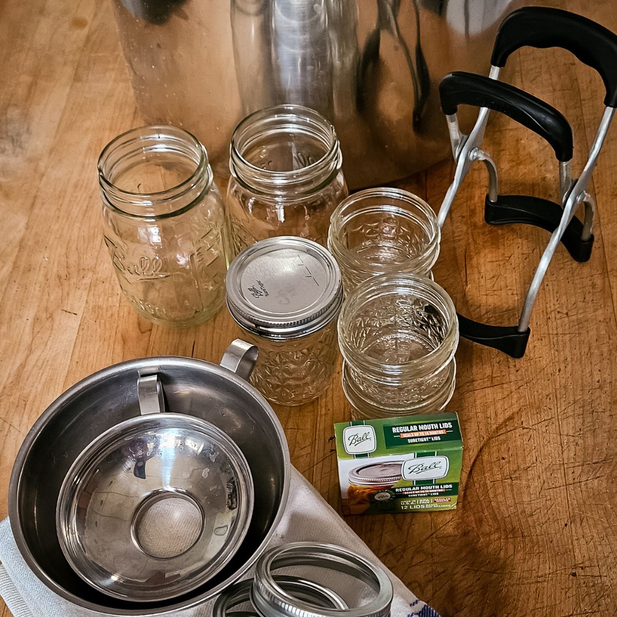 Canning supplies on a wooden surface, featuring a large metal pot, several empty jars with lids, a funnel, a towel, and jar lifter. The scene conveys readiness and organization.