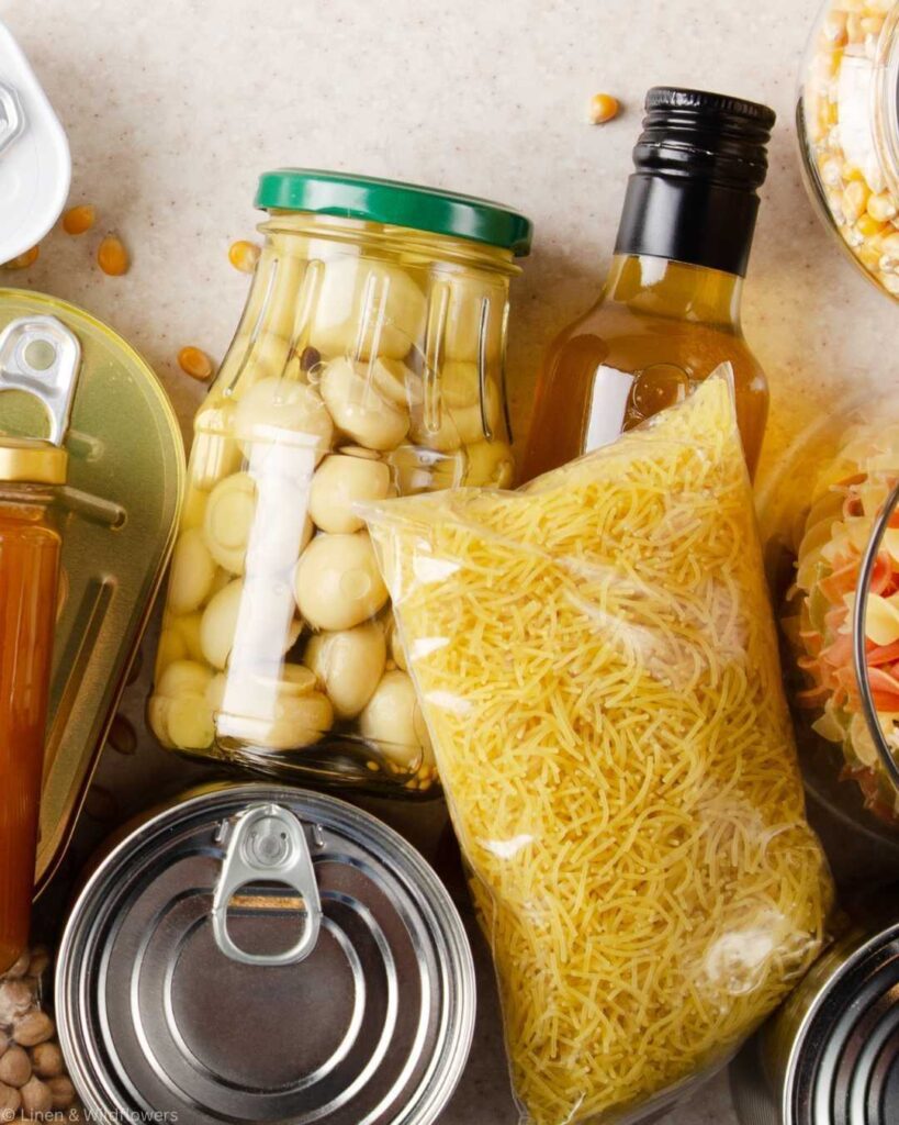 Close-up of assorted pantry items on a counter. Includes a jar of mushrooms, bottle of oil, bag of noodles, and canned goods, conveying a stocked kitchen.