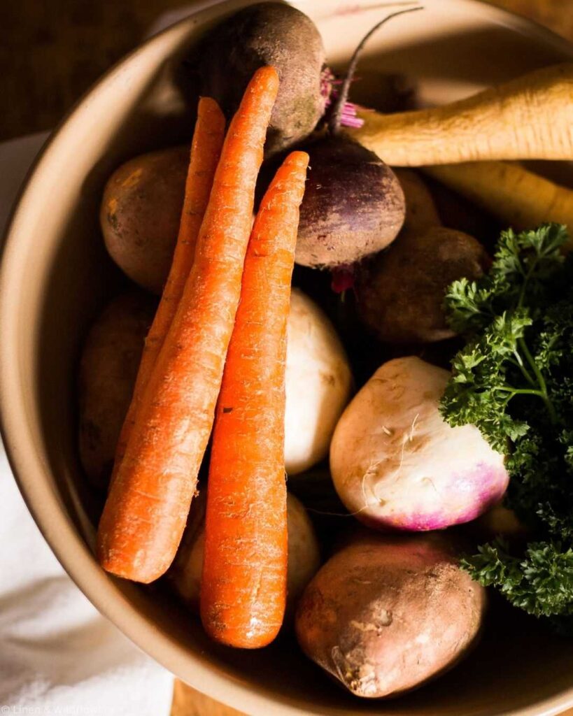 A bowl filled with fresh root vegetables, including vibrant orange carrots, beets, parsnips, and leafy parsley, creating a warm, earthy tone.