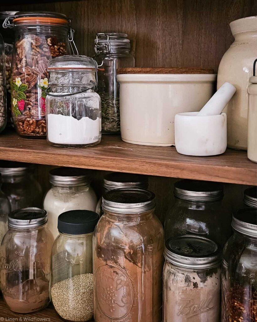 A wooden pantry shelf with various glass jars containing ingredients like pecans, seeds, quinoa, flour, and cocoa, alongside a cream crock and mortar.