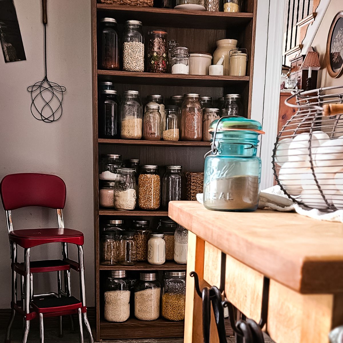 Kitchen scene with a wooden shelf filled with jars of grains and pasta. A countertop with hanging cast iron pans and a basket of eggs is in the foreground