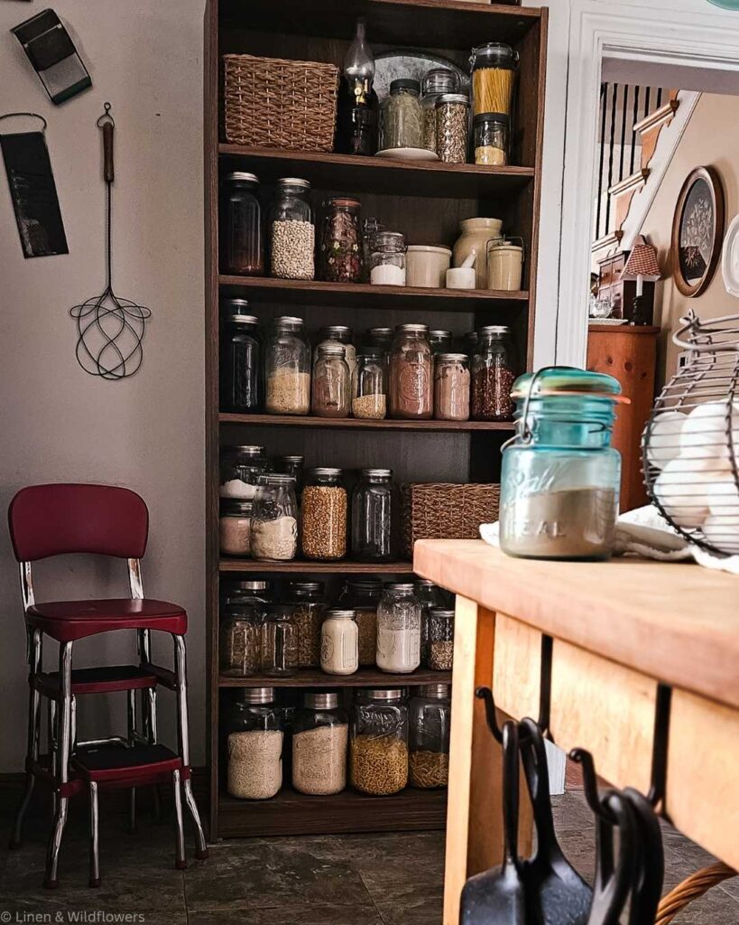 Kitchen scene with a wooden shelf filled with jars of grains and pasta. A countertop with hanging cast iron pans and a basket of eggs is in the foreground