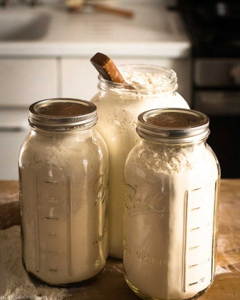 Three mason jars filled with white flour sit on a wooden table. Two have lids, and the third is open with a wooden scoop inside.