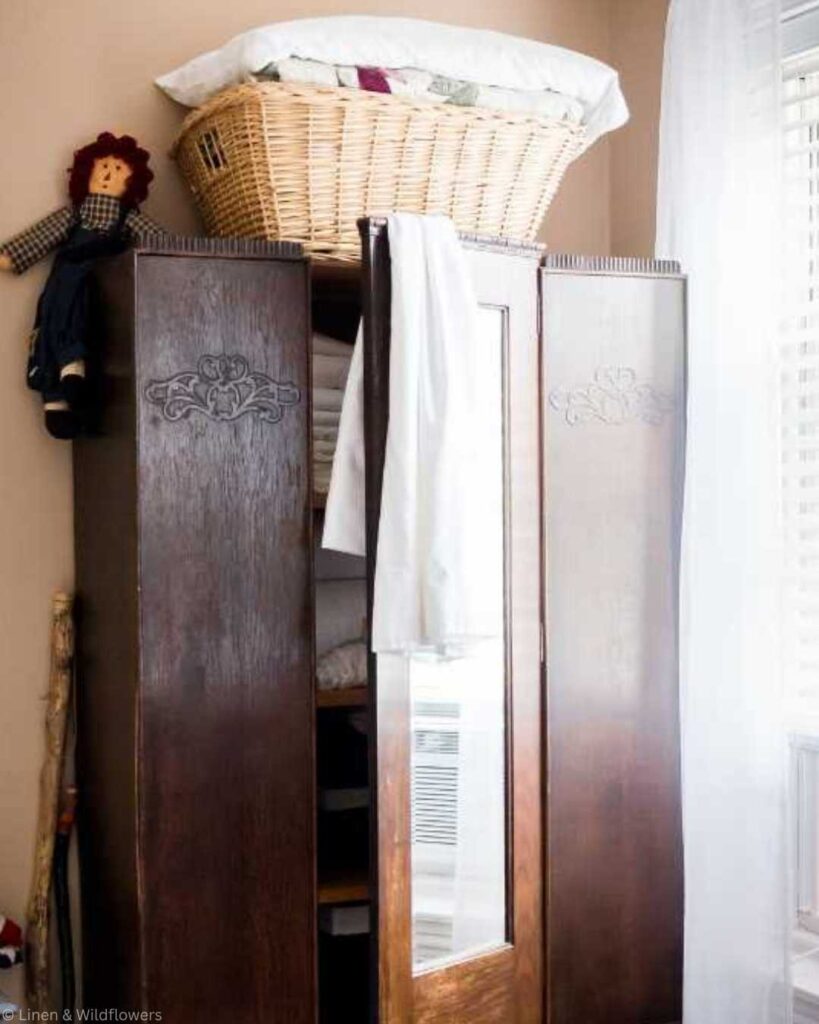 Vintage wooden wardrobe with a wicker basket on top, filled with quilts. A Raggedy Ann doll sits to the side,