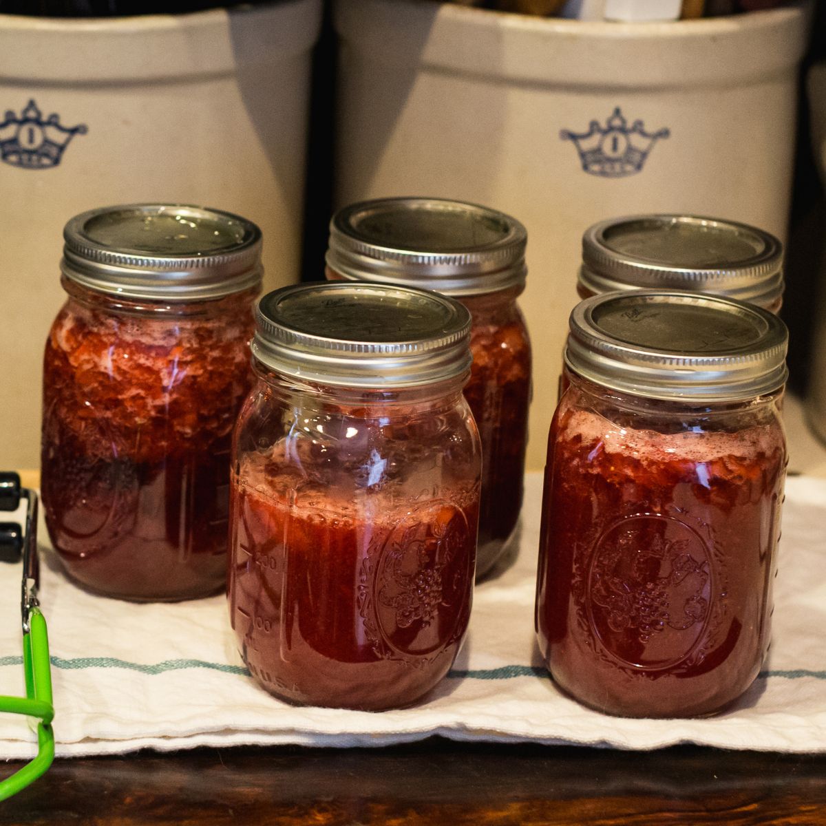Five glass jars filled with homemade strawberry jam rest on a cloth, radiating a warm, rustic charm. The background features ceramic crocks.