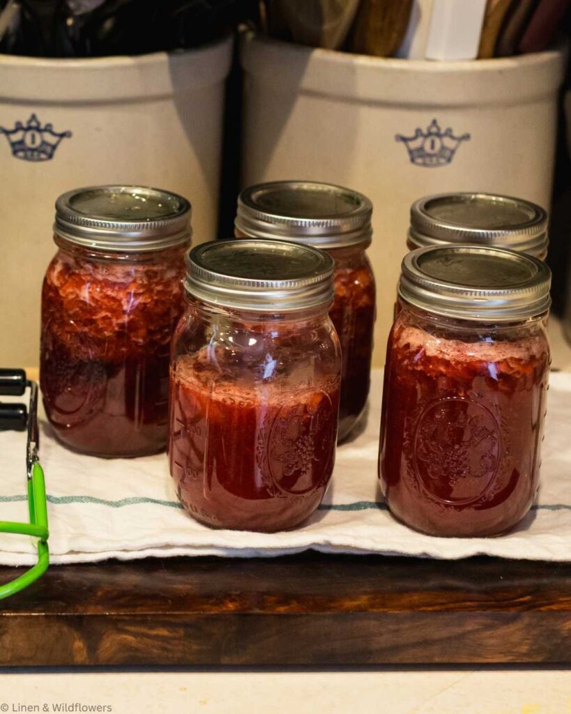 Five glass jars filled with homemade strawberry jam sit on a cloth-covered wooden surface. The jars have silver lids, conveying a cozy, homely kitchen setting.