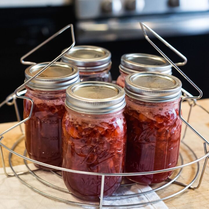 Five Mason jars filled with homemade red jam sit in a wire rack on a wooden countertop fresh out of the canner.