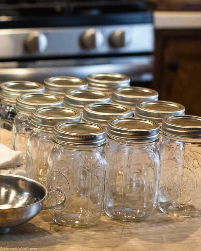 A kitchen scene with a stove in the background, displaying a large pot on a counter. Nearby are a stack of empty mason jars, a green jar lifter, a metal bowl, and dish towels, suggesting a canning preparation.