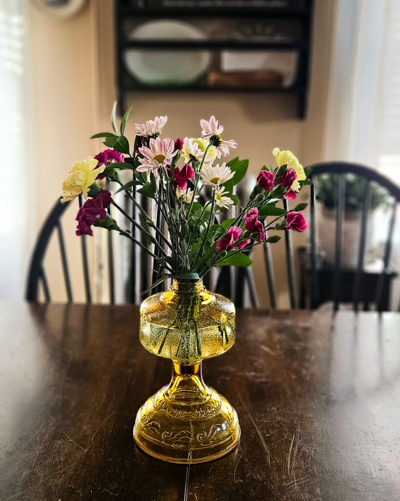 A bouquet of pink and cream flowers in a vintage amber glass vase on a dark wood dining table, with natural light filtering through the curtains.