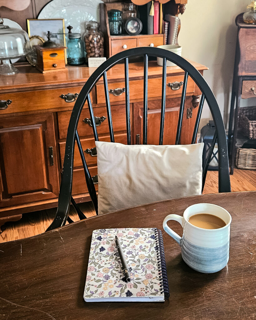 Cozy dining room setting with a wooden table, featuring a floral notebook and pen, and a blue mug of coffee. A cushioned chair and wooden cabinet add warmth.