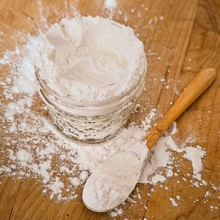 A small glass bowl filled with baking powder sits on a wooden surface. Flour is spilled around it, with a wooden spoon resting nearby, conveying a rustic baking scene.