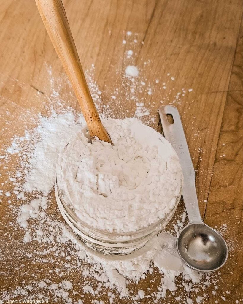 A wooden spoon in a glass jar filled with white baking soda or flour on a wooden surface. A metal measuring spoon lies beside it, with scattered powder around.