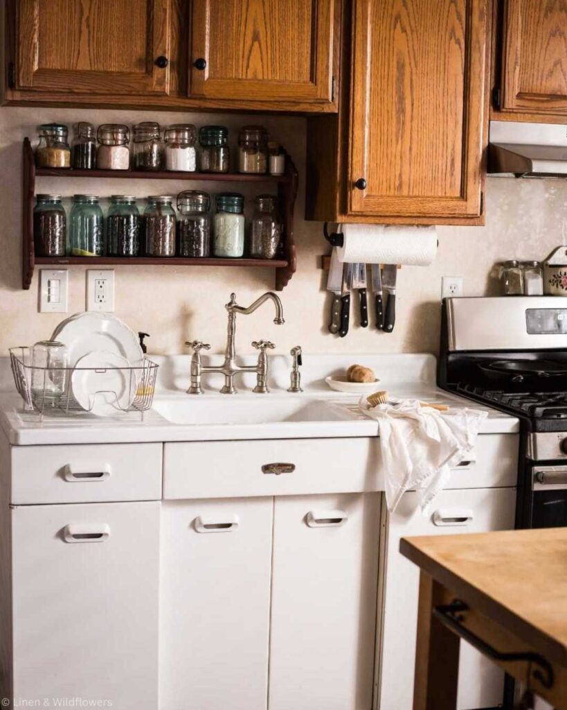 Cozy kitchen with wooden cabinets, white sink, and vintage faucet. Mason jars are on shelves, and knives are beside a stove.
