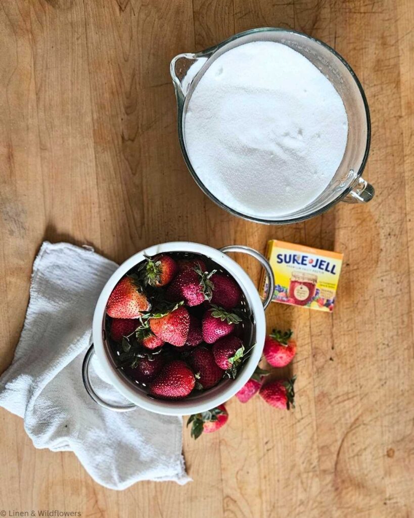 Bowl of fresh strawberries beside a container of sugar and a box of Sure-Jell on a wooden countertop, conveying a homemade jam-making vibe.