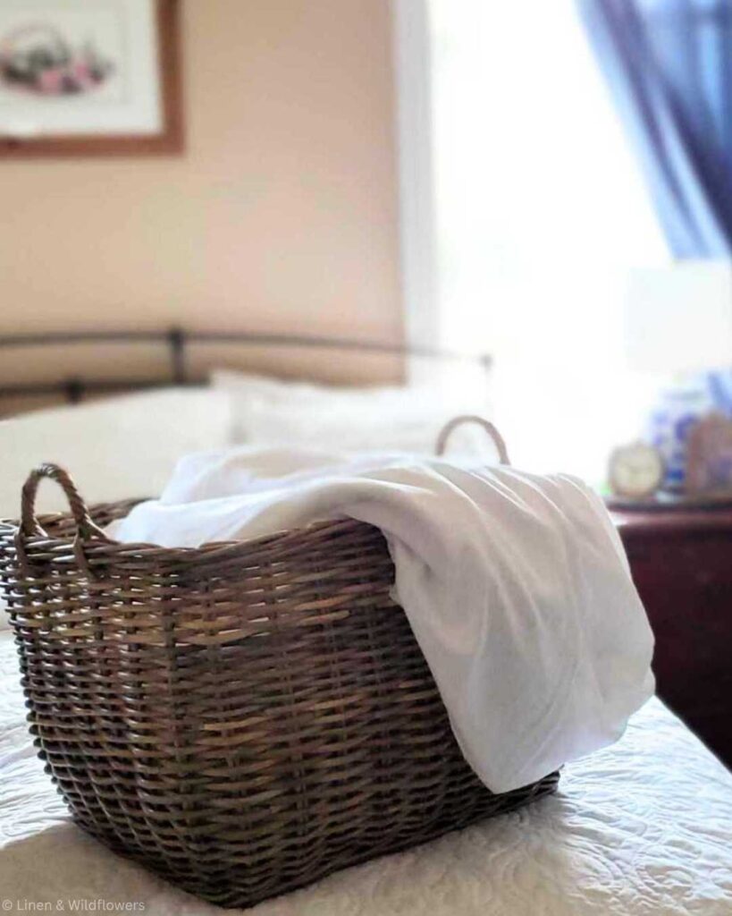 A wicker basket with white linens sits on a quilted bed in a cozy, sunlit bedroom. The scene conveys a peaceful, homely atmosphere.