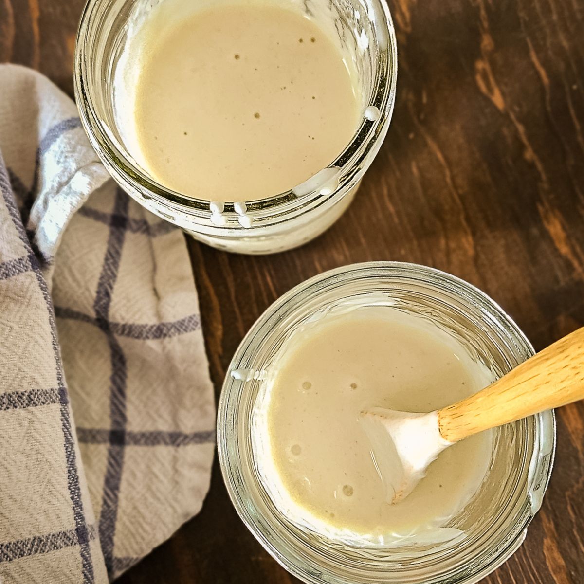 Two glass jars filled with sourdough discard sit on a wooden table. A wooden spoon rests in one jar, and a checkered cloth lies beside them, creating a cozy and inviting scene.