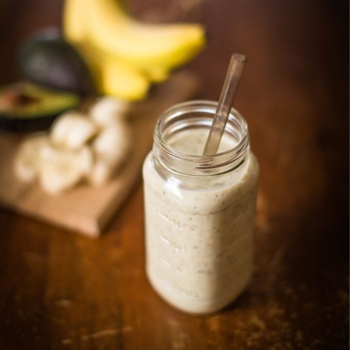 A refreshing green smoothie made with avocado and banana, served in a mason jar with a glass straw, with blurred fruit ingredients on a rustic wooden surface.