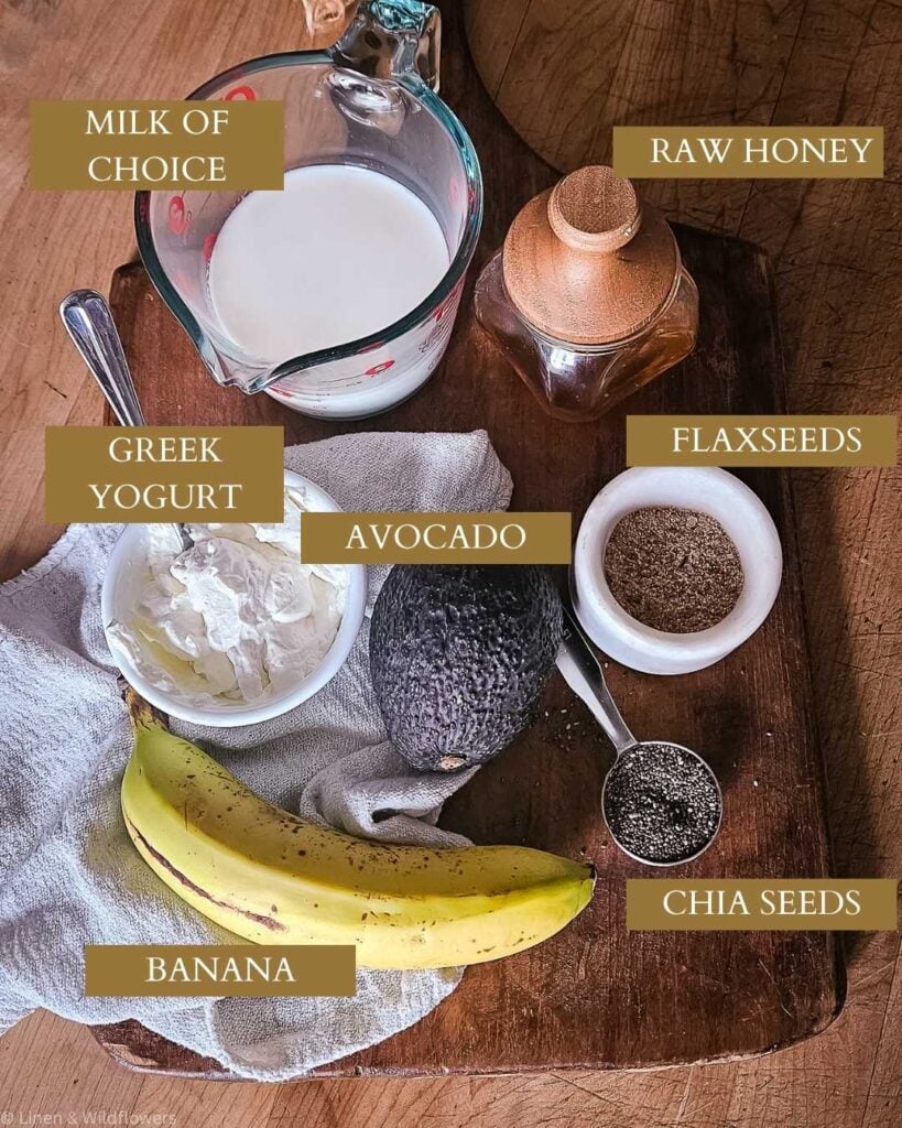 Ingredients for a smoothie on a wooden board: milk in a jug, raw honey in a jar, flaxseeds, chia seeds, an avocado, Greek yogurt, and a banana.