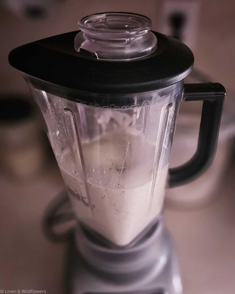 A blender with a black lid is half-filled with a creamy banana avocado smoothie. The background is softly blurred, creating a cozy kitchen ambiance.