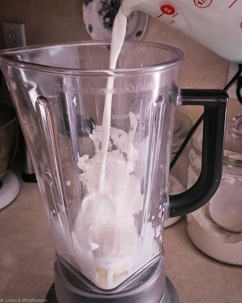 A blender on a kitchen counter is filled with smoothie ingredients, and milk is being poured in. The scene conveys a cozy, homemade feel, suggesting smoothie preparation.