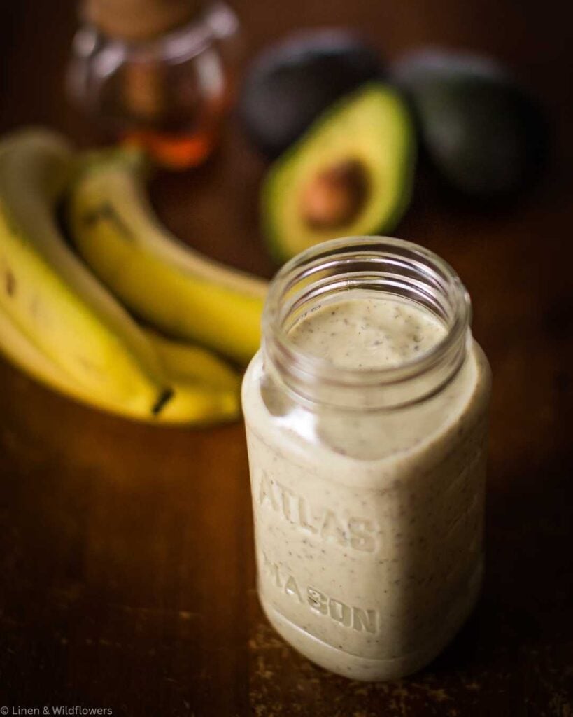 A thick avocado banana smoothie in an Atlas mason jar with fresh bananas, halved avocado, and a jar of honey softly blurred in the background.