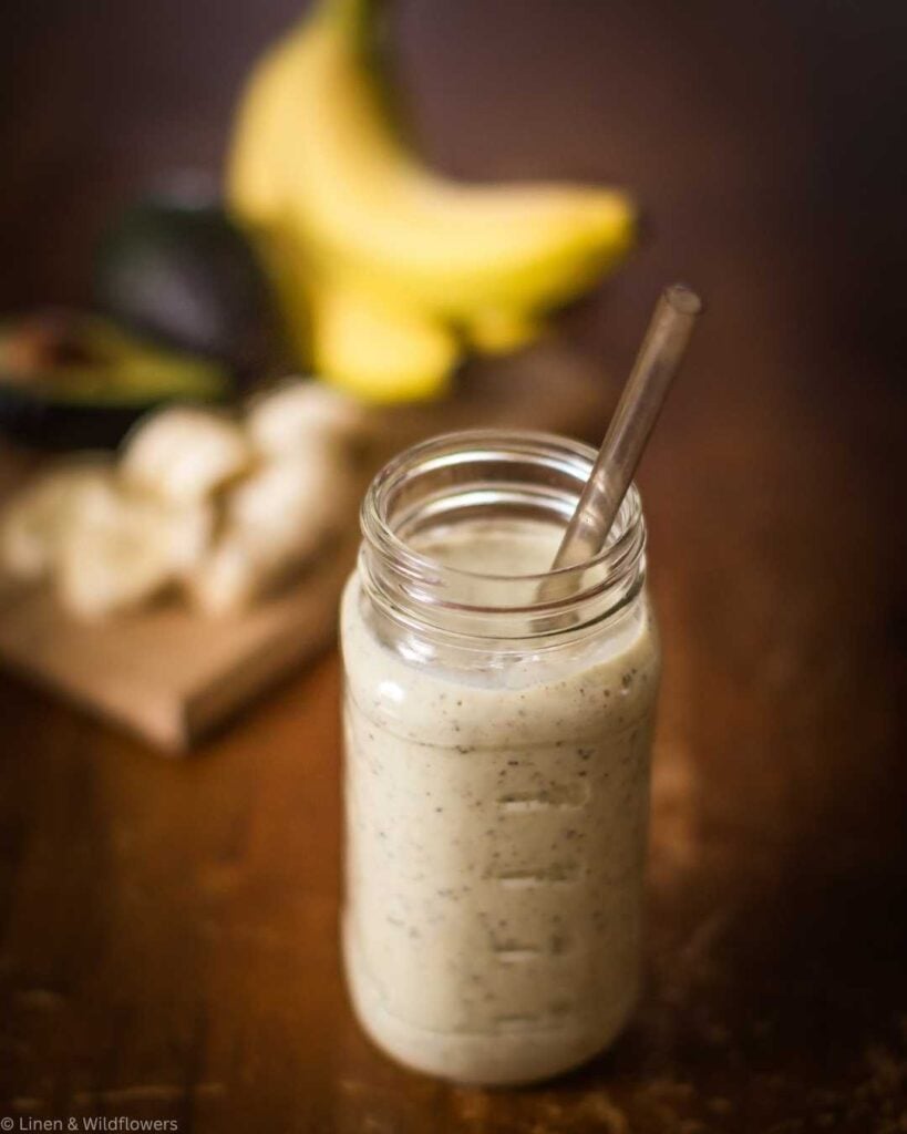 A refreshing green smoothie made with avocado and banana, served in a mason jar with a glass straw, with blurred fruit ingredients on a rustic wooden surface.