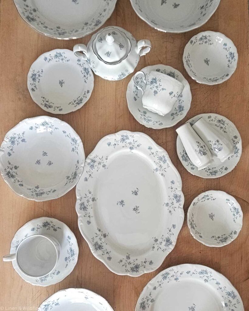 A collection of elegant white china with delicate blue floral patterns arranged on a wooden table. Includes plates, bowls, a teapot, and cups, conveying a vintage feel.