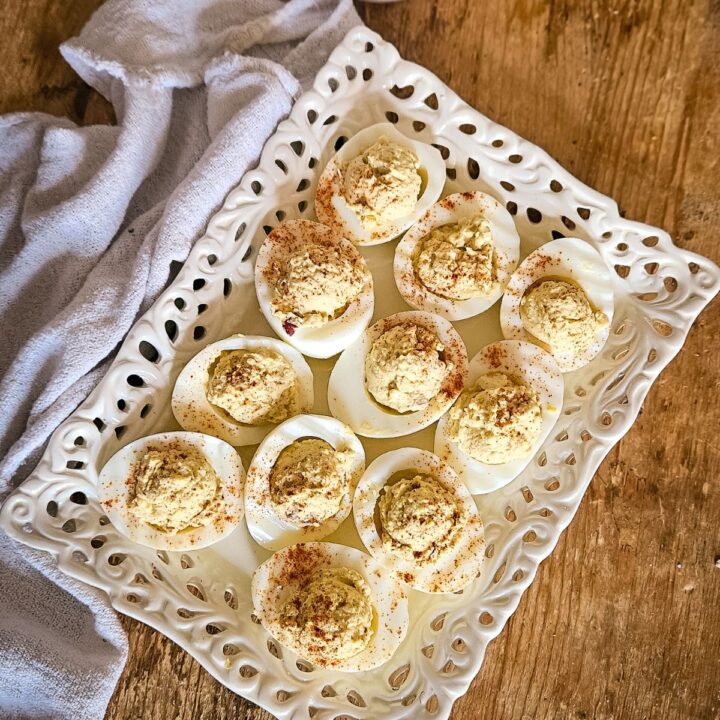 Deviled eggs topped with paprika on a decorative white platter. Wooden table surface and a soft, folded cloth napkin add a rustic touch.
