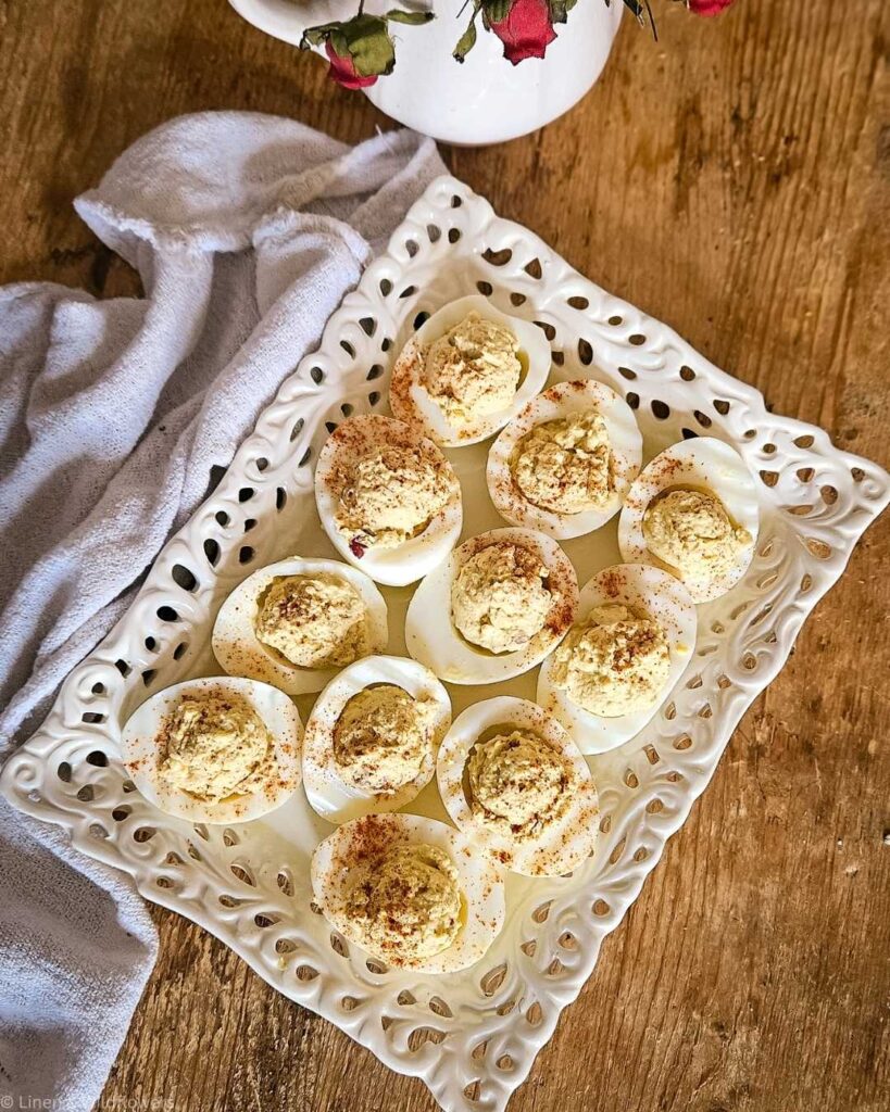 Deviled eggs sprinkled with paprika sit on a decorative white platter. A textured cloth is nearby, beside a vase with red roses, on a wooden table.