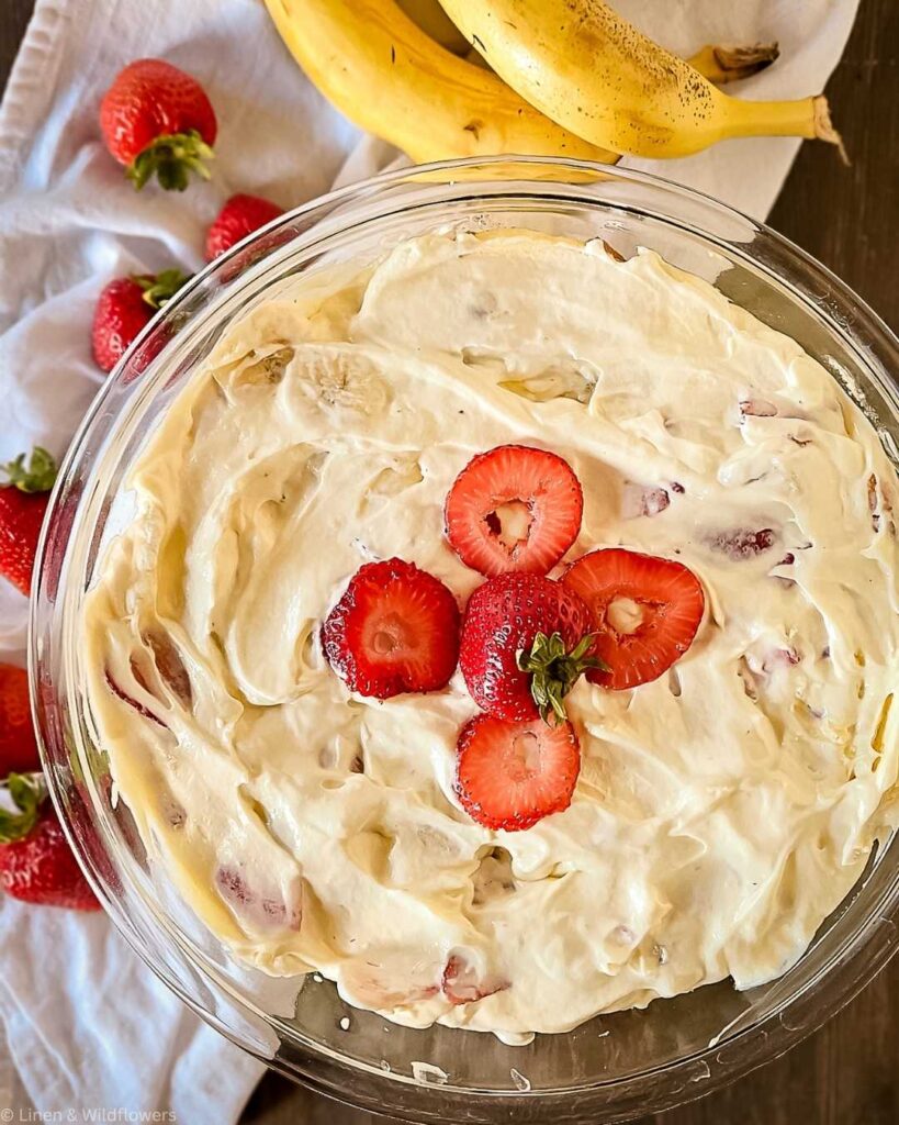 A glass bowl filled with creamy banana strawberry dessert, topped with fresh strawberry slices. Bananas and whole strawberries are on a white cloth nearby.