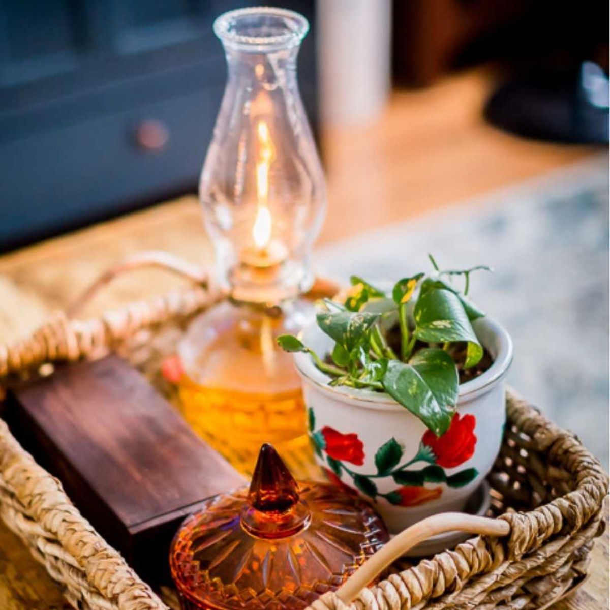 A wicker basket holds a lit oil lamp, a potted plant with green leaves in a colorful floral pot, a wooden box, and an amber glass jar.