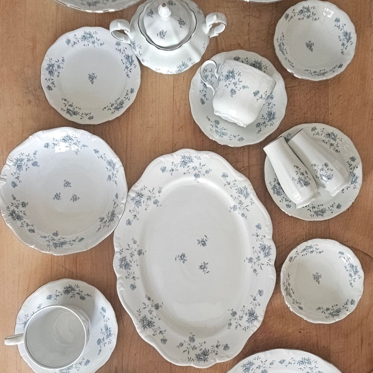 A collection of elegant white china with delicate blue floral patterns arranged on a wooden table. Includes plates, bowls, a teapot, and cups, conveying a vintage feel.