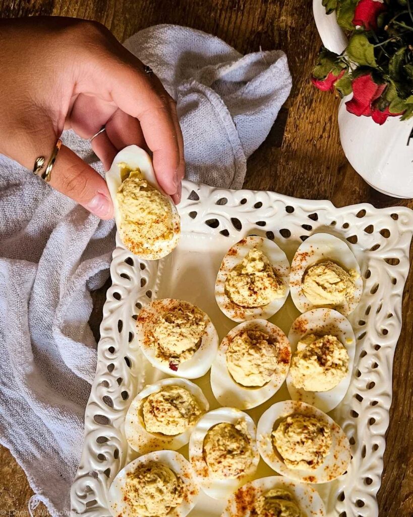 A hand holding a deviled egg over a white ornate tray filled with more deviled eggs, sprinkled with paprika. A white cloth and flowers are beside the tray.