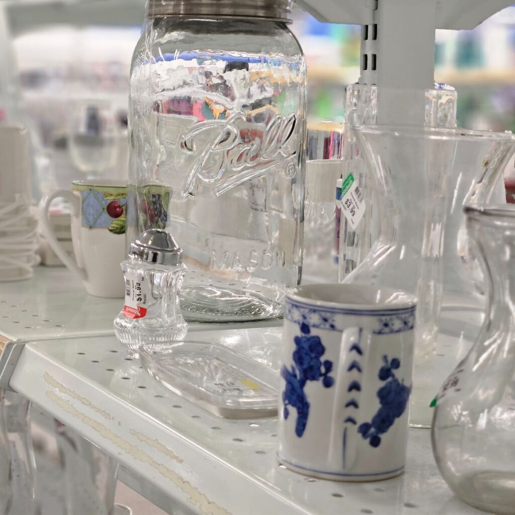 Shelves display various glassware, including a large mason jar, clear salt shaker, and a white mug with blue floral design, evoking a vintage feel.