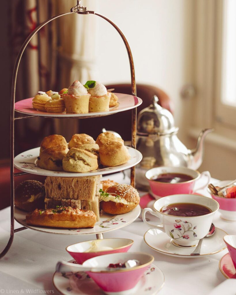 Three-tiered stand with assorted pastries, scones, and sandwiches on a table. Tea set with floral cups and a silver teapot, conveying elegance and warmth.