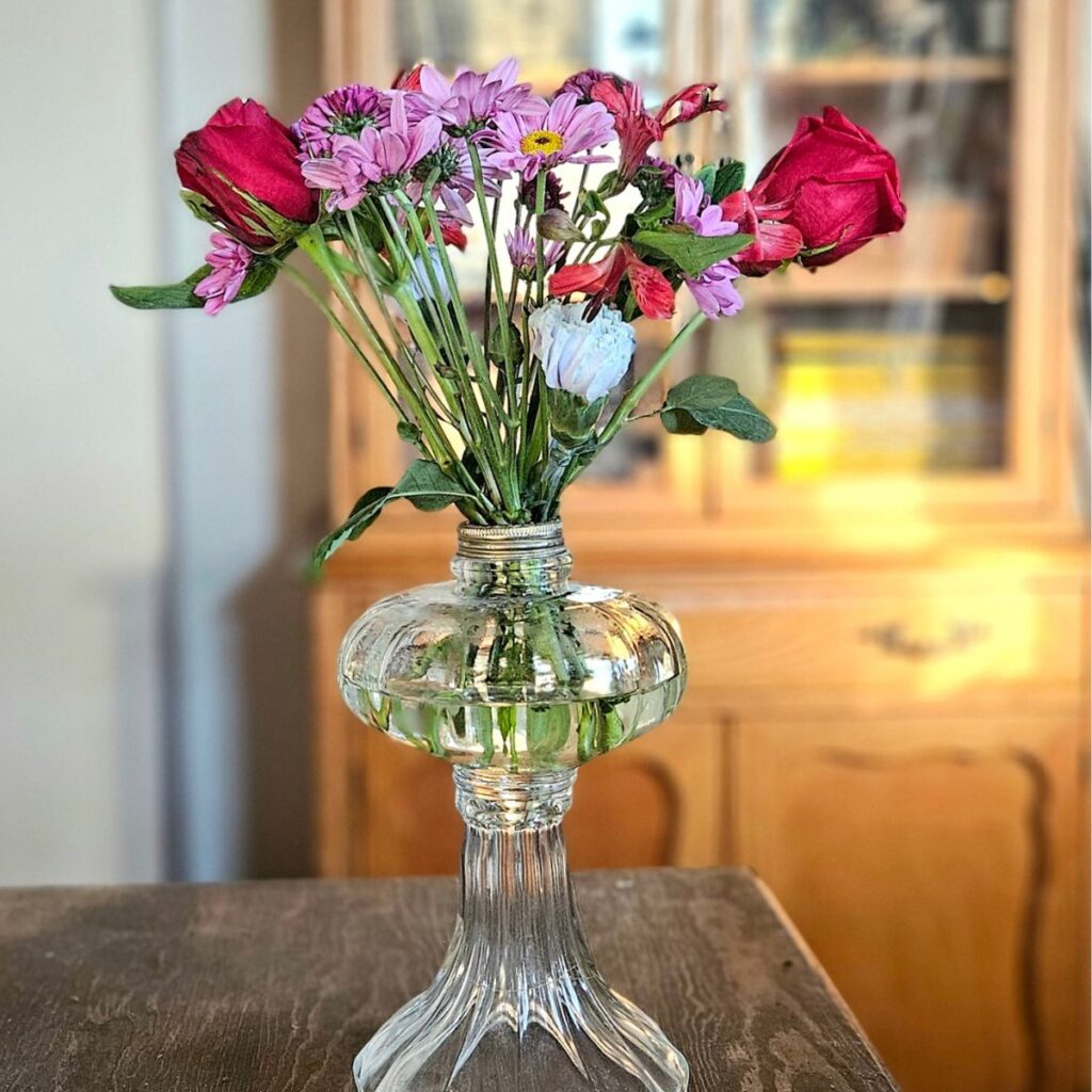 A clear glass vase holds red roses and purple daisies on a dark wooden table. Soft, warm light creates a cozy atmosphere with a wooden cabinet in the background.