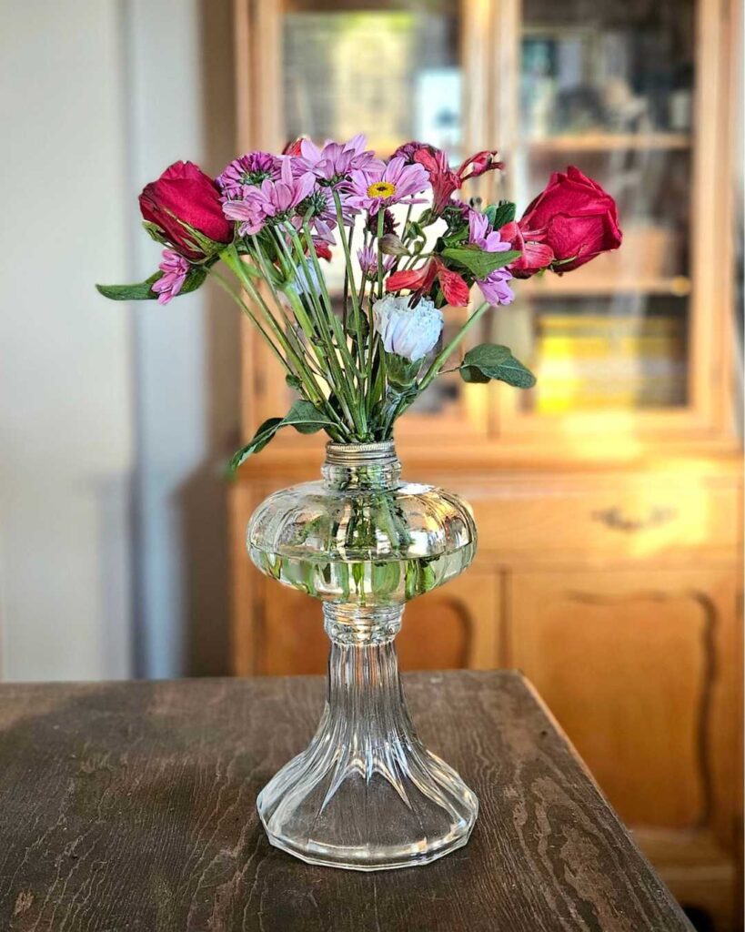 A clear glass vase holds red roses and purple daisies on a dark wooden table. Soft, warm light creates a cozy atmosphere with a wooden cabinet in the background.