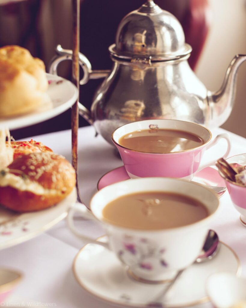 Elegant afternoon tea setting with pink teacups, a silver teapot, and a tiered tray of pastries. The atmosphere is warm and inviting.