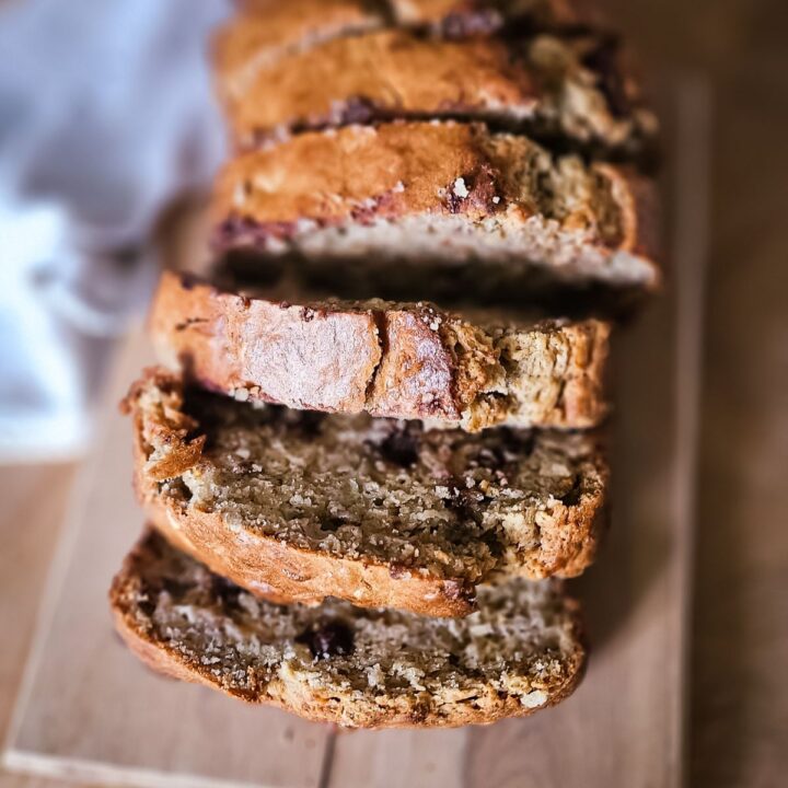 Sliced banana bread on a wooden board, showing a golden-brown crust and moist interior with chocolate chips. A cozy, homemade feel is conveyed.