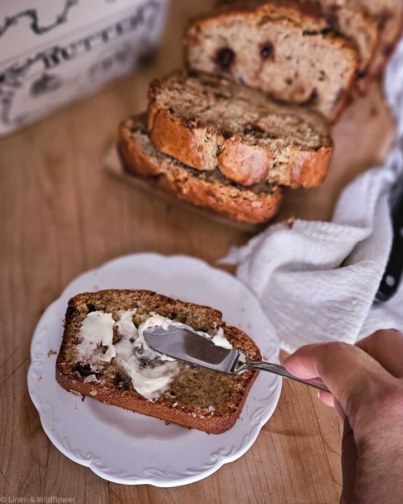 A hand spreads creamy butter on a slice of banana bread, placed on a white plate. More slices are stacked nearby, creating a cozy, homemade feel.