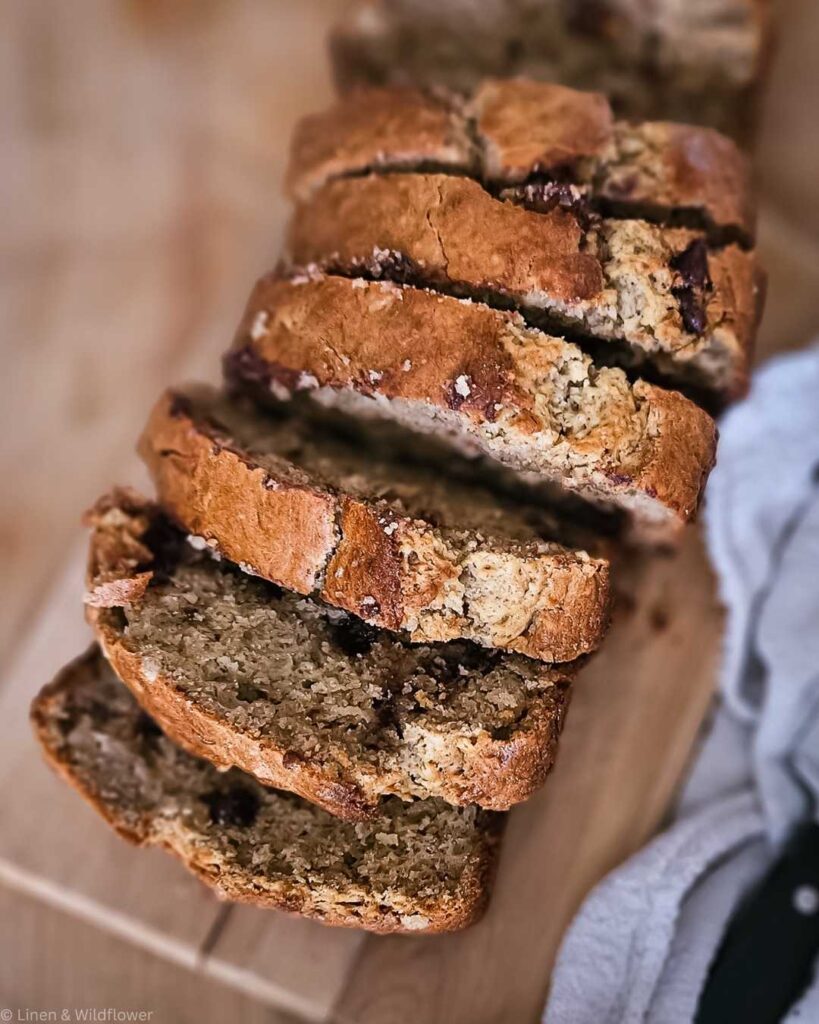 Sliced banana bread on a wooden board, showing a golden-brown crust and moist interior with chocolate chips. A cozy, homemade feel is conveyed.