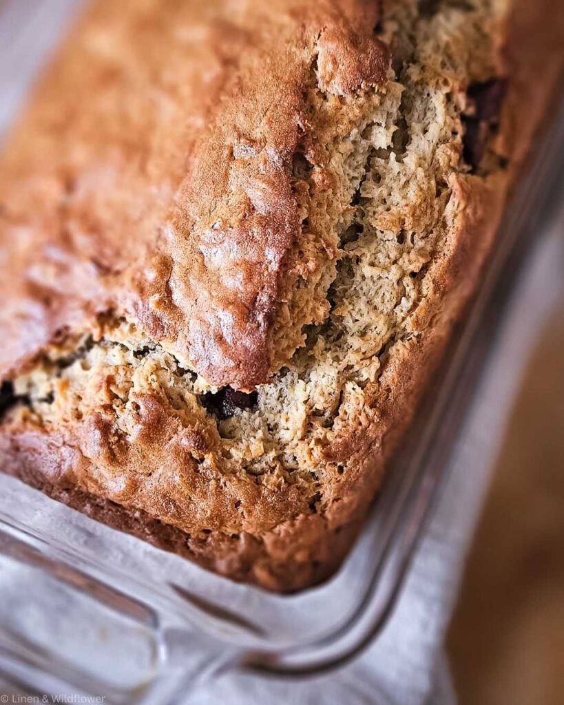 Close-up of a golden-brown, freshly baked banana bread in a glass dish. The texture is crumbly and inviting, evoking warmth and comfort.