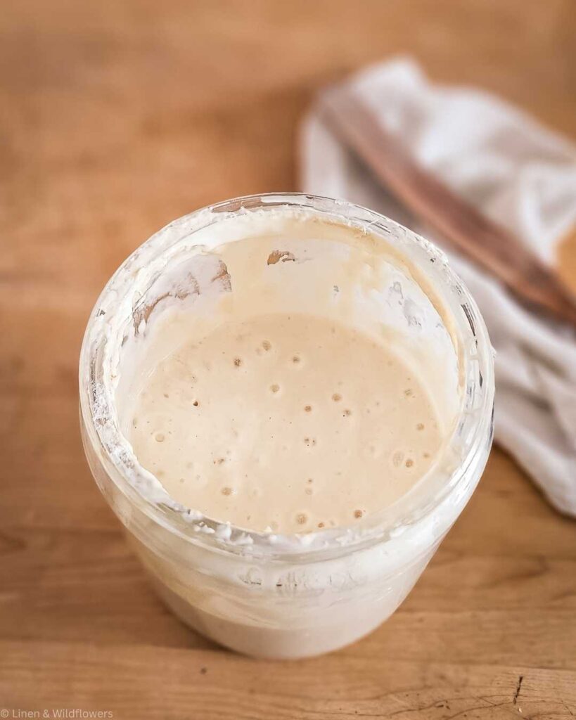 A glass jar filled with creamy sourdough starter sits on a wooden surface. The dough is bubbly and active, conveying freshness and homemade warmth.