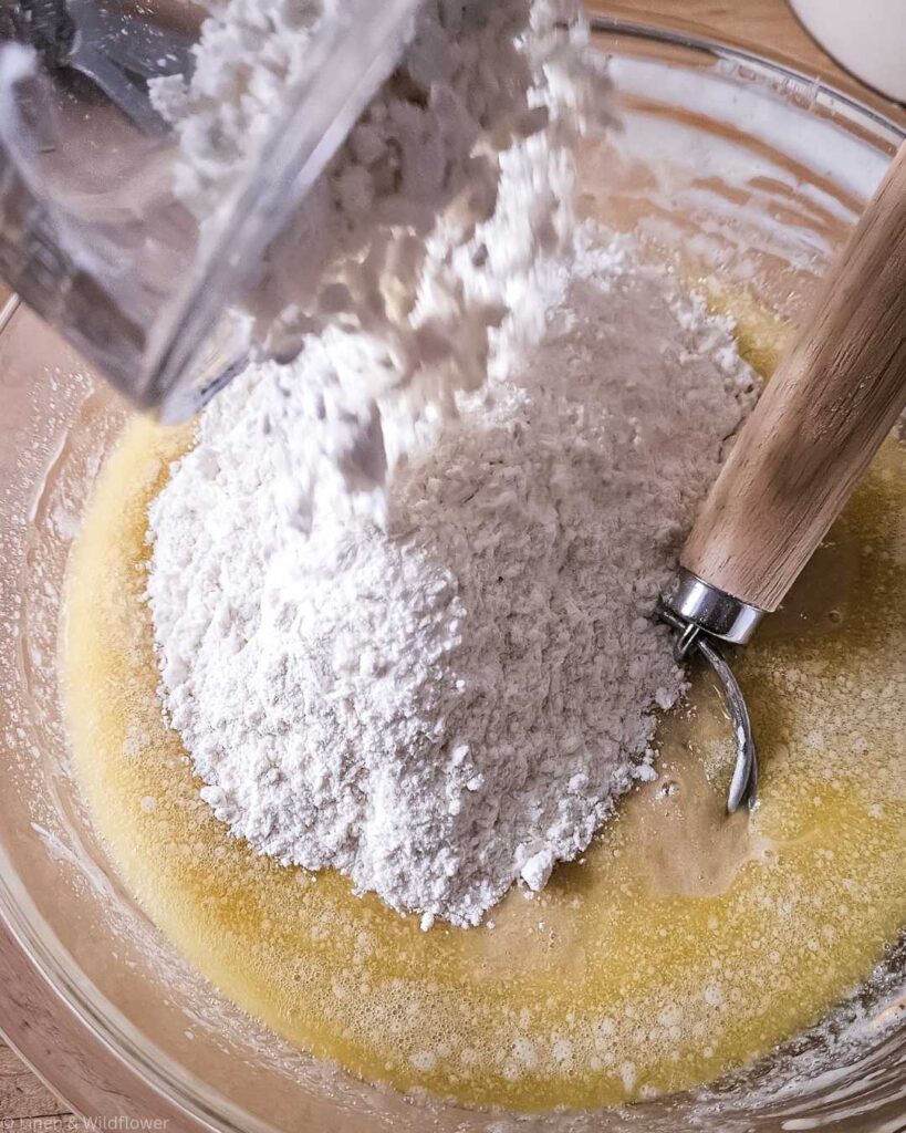 Flour is being poured into a bowl with a yellow liquid mixture, possibly eggs and butter. A wooden-handled dough whisk rests in the mix, suggesting baking preparation.